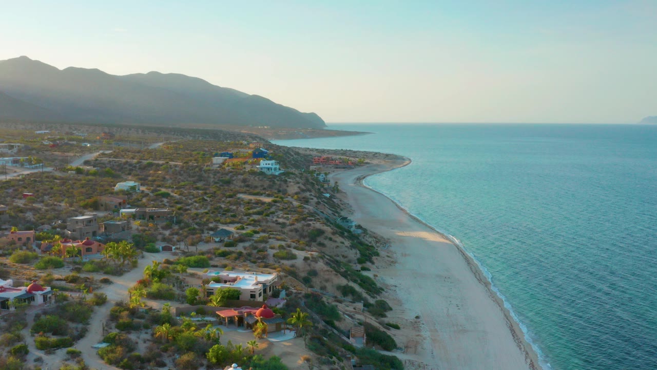 Aerial view of El Sargento La Ventana, Mexico, with turquoise water beside a pale curving beach, scattered homes and scrubland in the foreground, mountains in the distance, and no visible people.