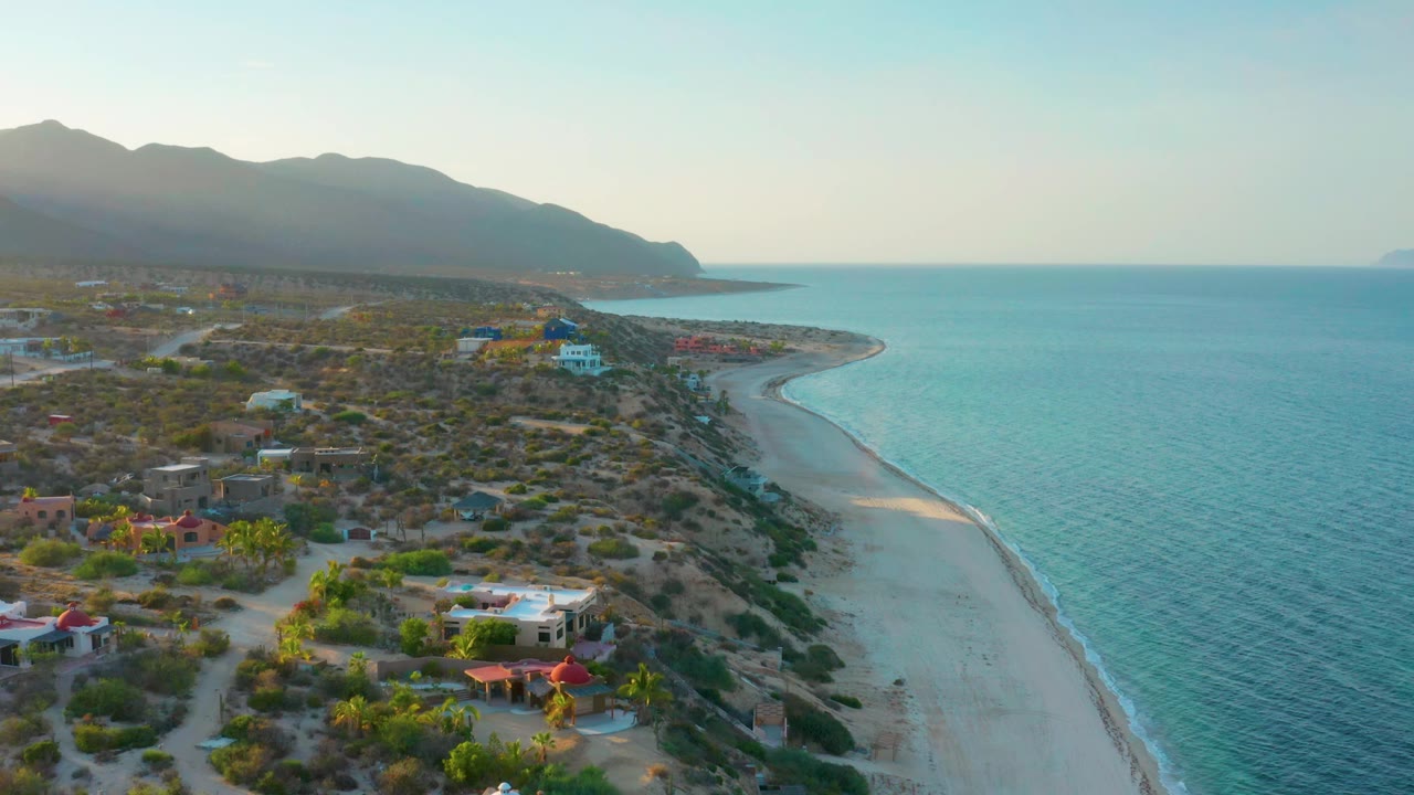 Aerial frame over El Sargento La Ventana showing coastal houses and desert vegetation to the left, a narrow pale beach along the center, blue ocean to the right, and no visible people.