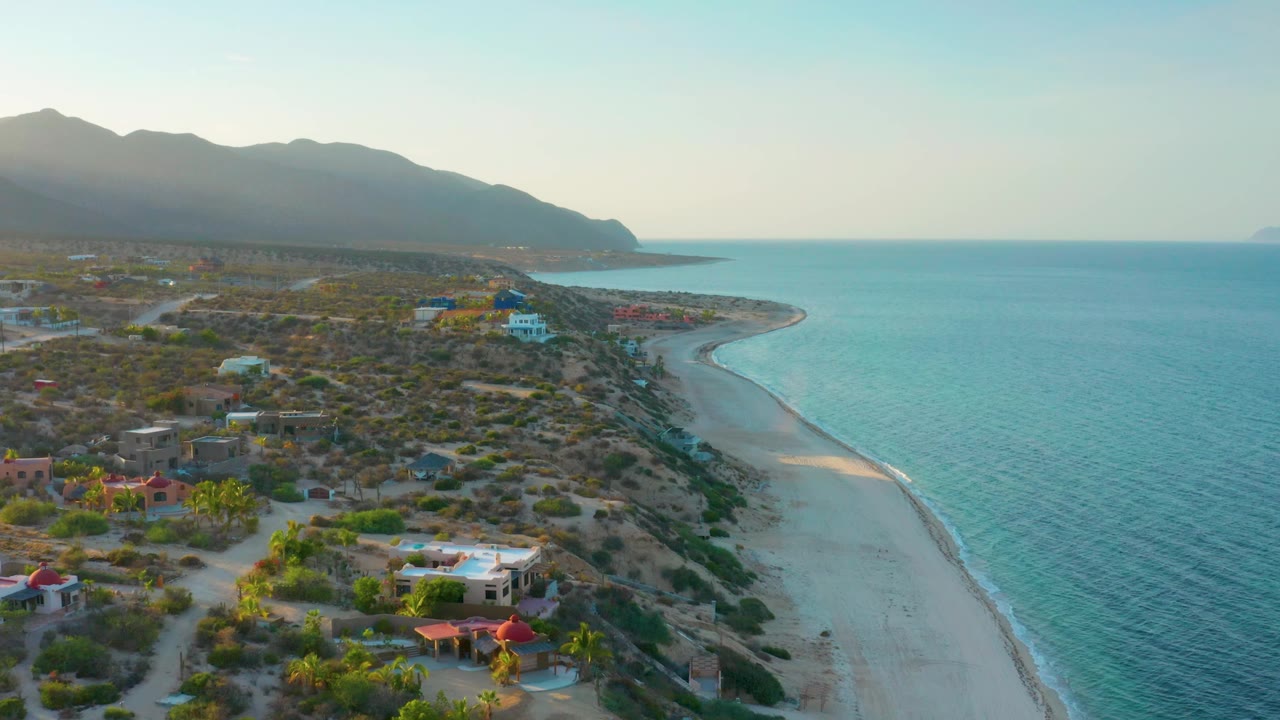 Wide drone view of El Sargento La Ventana with a bright turquoise sea filling the right side, beach and rocky edge below, colorful low buildings on the left, distant mountains, and no visible people.