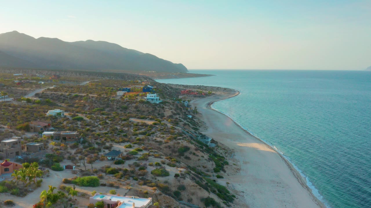 Opening aerial view of El Sargento La Ventana, Mexico with blue sea on the right, pale sand beach curving below, desert scrub and small buildings inland, mountains in haze, and no visible people.