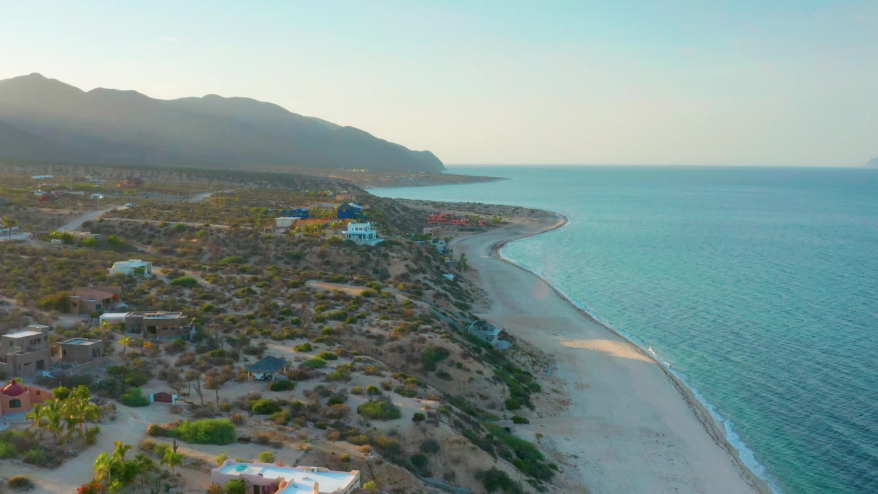 Elevated drone view of beach, turquoise water, coastal homes, scrubland, and distant mountains.