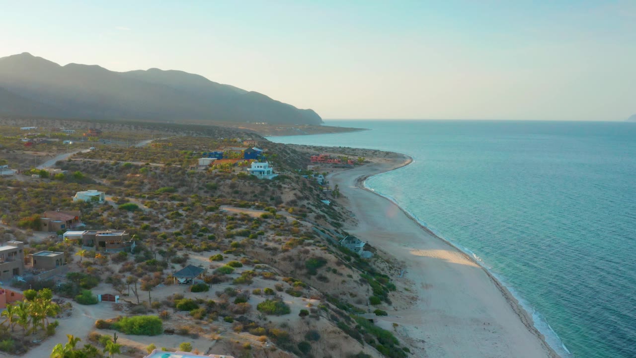 A wide aerial frame in El Sargento La Ventana, Mexico showing the shoreline bending toward a point, bright blue sea to the right, sparse coastal buildings to the left, mountain ridge behind, and no visible people.