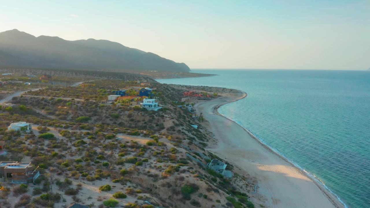 Wide drone view over El Sargento La Ventana, Mexico, with dry brush and houses on the dunes, a narrow beach below, turquoise sea to the right, mountain silhouettes beyond, and no visible people.