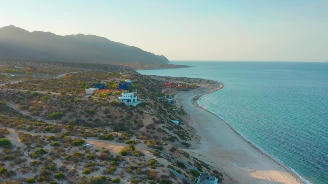 Opening aerial frame over El Sargento La Ventana, Mexico, with scrub-covered land and scattered houses at left, a curving pale beach beside blue-green water at right, hazy mountains beyond, and no visible people.