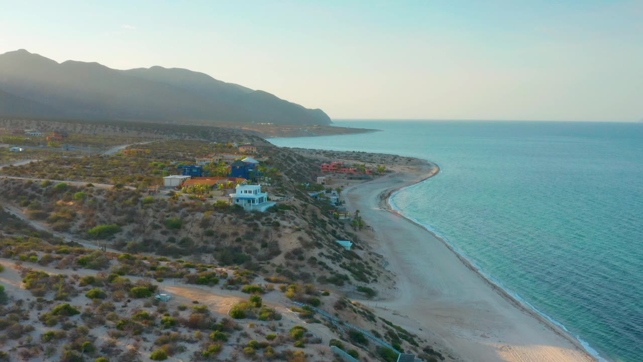 Drone frame over El Sargento La Ventana, Mexico, with the coastline running from lower center toward the distant point, scattered homes across dry terrain, turquoise water filling the right side, and no visible people.