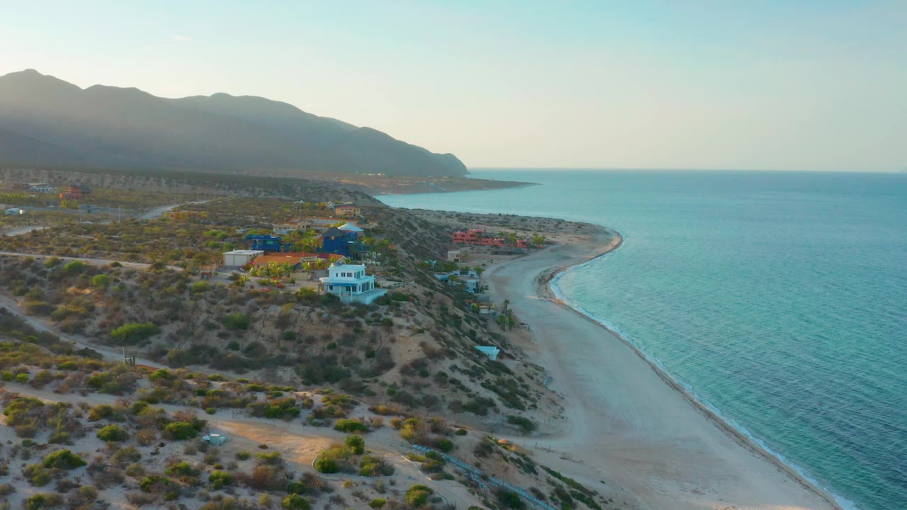Aerial coastline with a sandy beach, blue water, scrubland, houses, and hazy mountains.