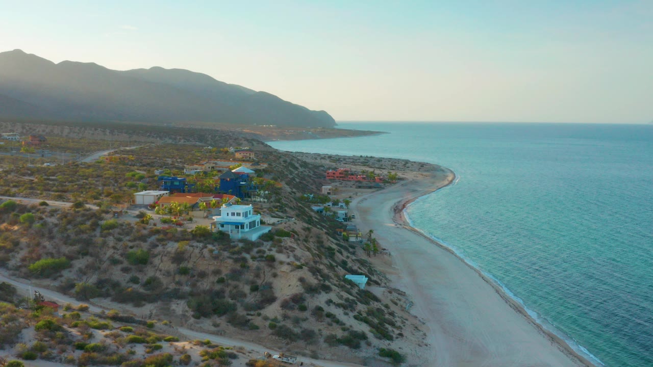 Aerial view in El Sargento La Ventana, Mexico with beach-side buildings on scrubby terrain, a curved sandy shore, blue ocean, distant mountains, and no visible people.
