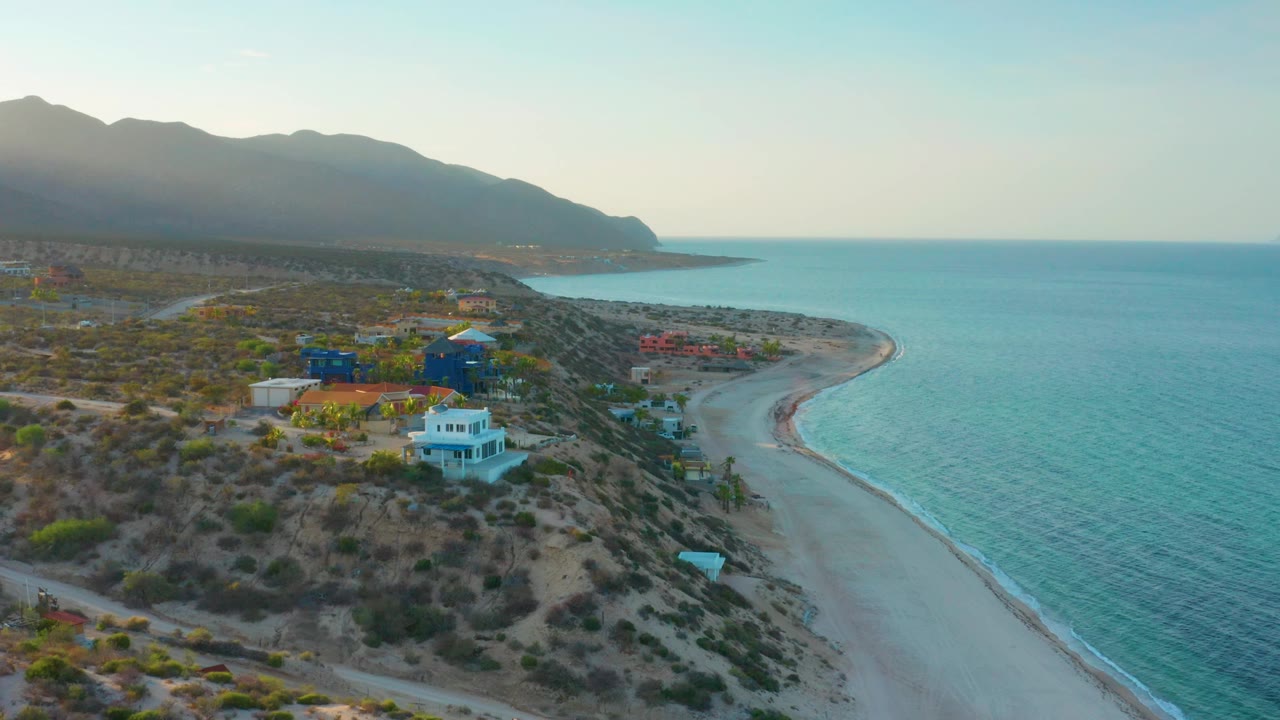 Wide aerial frame in El Sargento La Ventana, Mexico with clustered beach-side buildings, a pale sand curve, calm blue sea, mountain silhouettes, and no visible people.