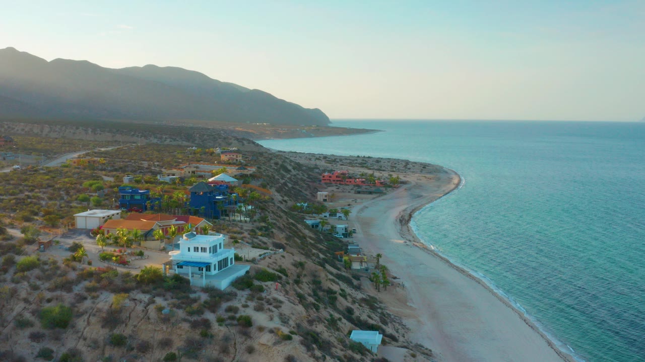 Opening aerial frame over El Sargento La Ventana, Mexico, with a white beach house in the lower foreground, colorful homes behind it, a curved sandy shoreline, blue ocean, distant mountains, and no visible people.