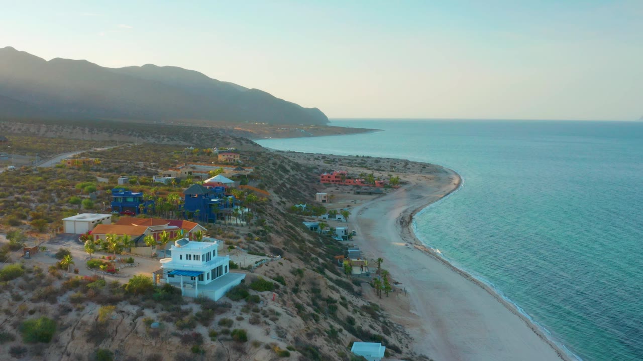 Mid-clip aerial view over El Sargento La Ventana with the beach houses shifted lower in frame, a longer sweep of curved sand visible, teal water filling the right side, hazy mountains beyond, and no visible people.