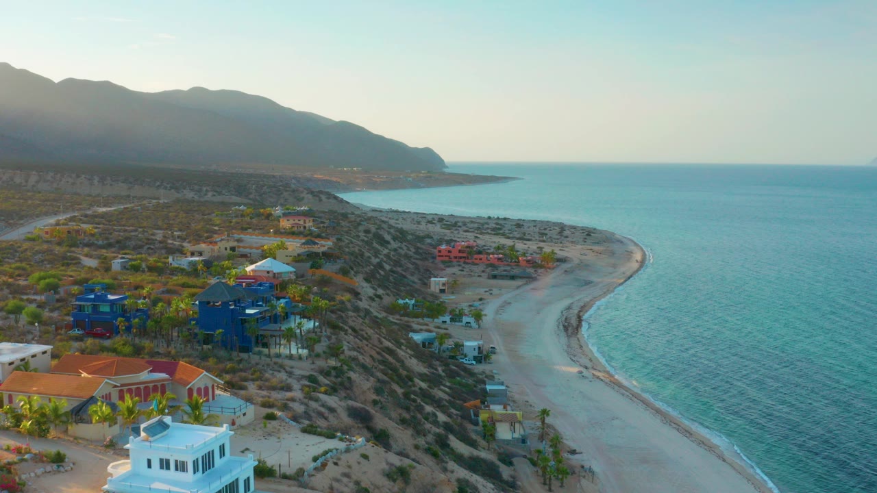 Aerial frame over El Sargento La Ventana, Mexico, showing colorful coastal homes, a pale curved beach, blue ocean, hazy mountains, and no visible people.