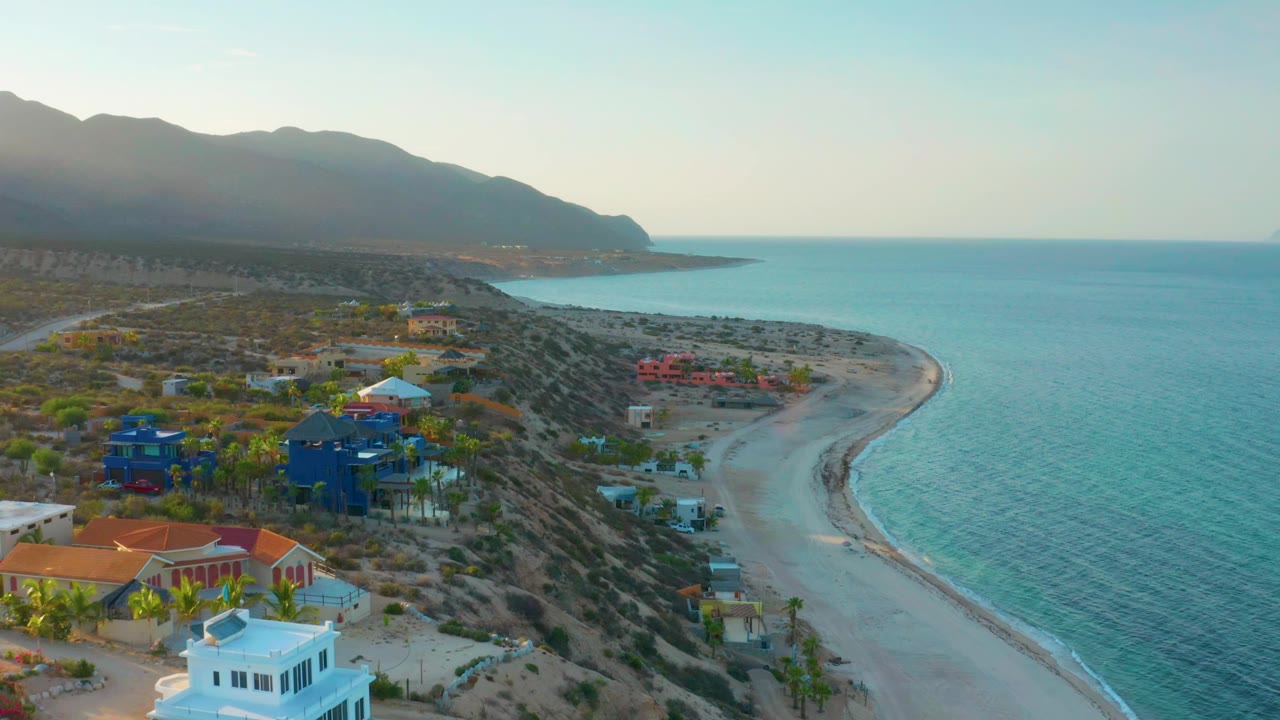 Wide drone view in El Sargento La Ventana, Mexico, with the beach arc on the right, clustered homes on the left, calm blue water, distant mountains, and no visible people.