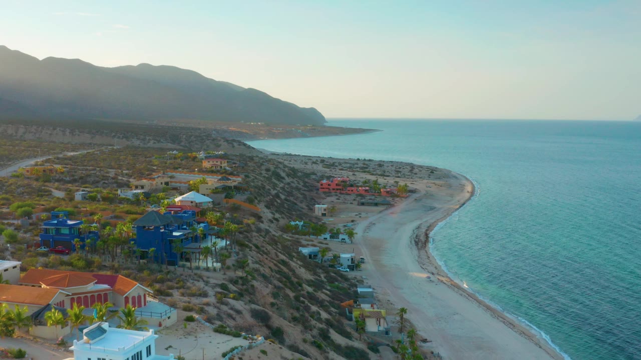 Aerial view over El Sargento La Ventana, Mexico, with the empty sand beach curving below dark mountains, blue-green ocean to the right, homes to the left, and no visible people.