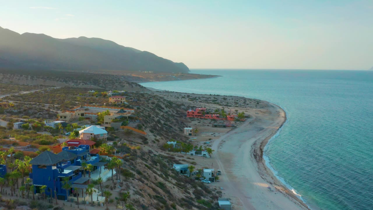 High aerial frame in El Sargento La Ventana, Mexico, with colorful foreground houses, dry scrubland, a narrow beach curve, calm blue water, distant mountains, and no visible people.