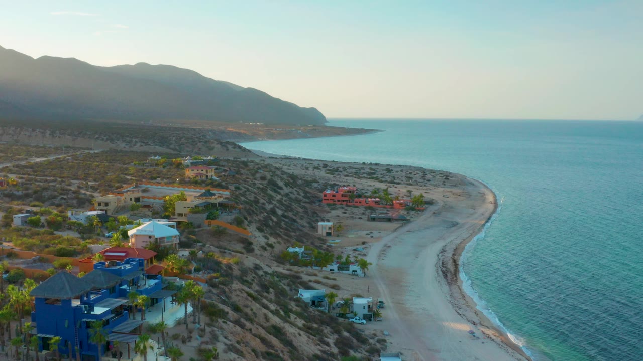 Aerial frame over El Sargento La Ventana, Mexico, showing a small coastal neighborhood, pale sand, a sweeping shoreline, blue sea, hazy mountains, and no visible people.