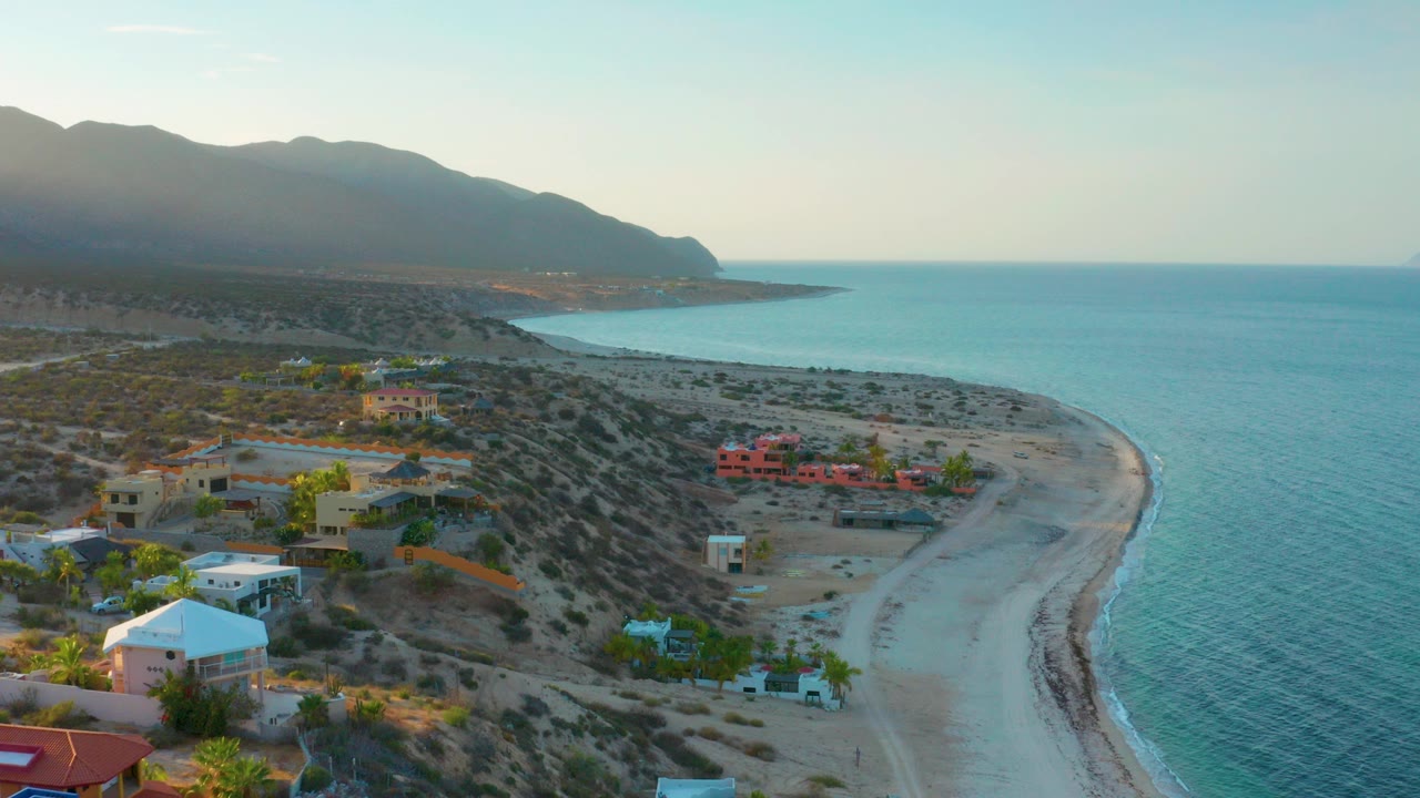 Aerial view over El Sargento La Ventana, Mexico with beach homes in the foreground, a curved sandy shore, turquoise water, distant mountains, and no visible people.
