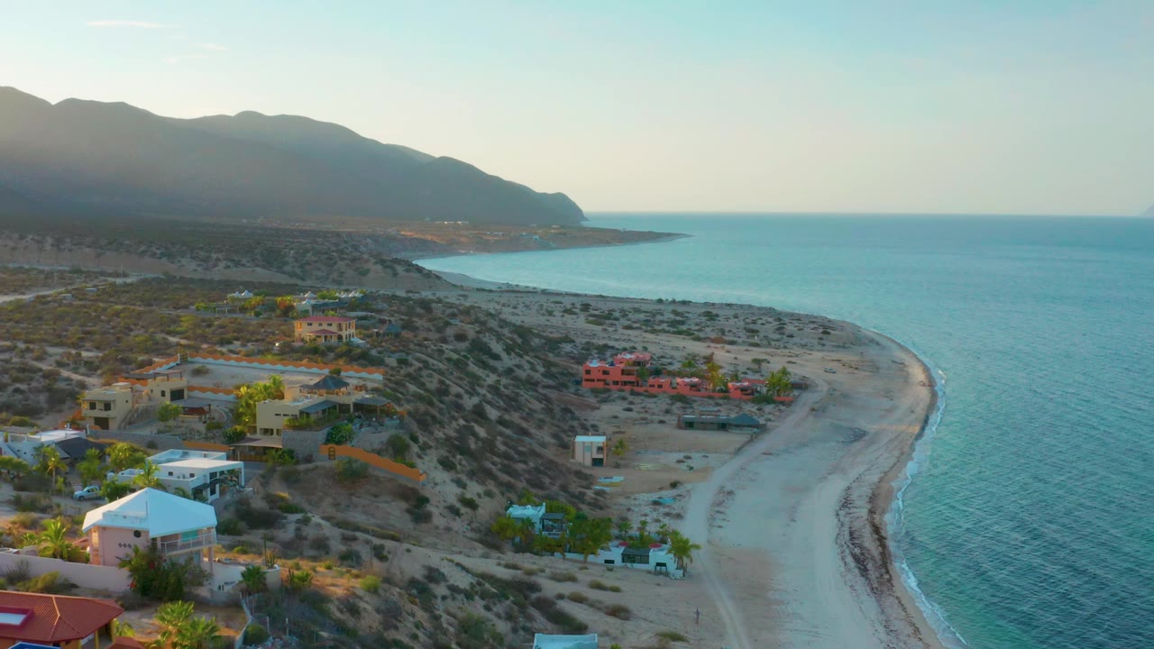High drone view in El Sargento La Ventana, Mexico showing scattered beachside homes, scrub-covered lots, a pale sand curve, blue-green water, mountains, and no visible people.