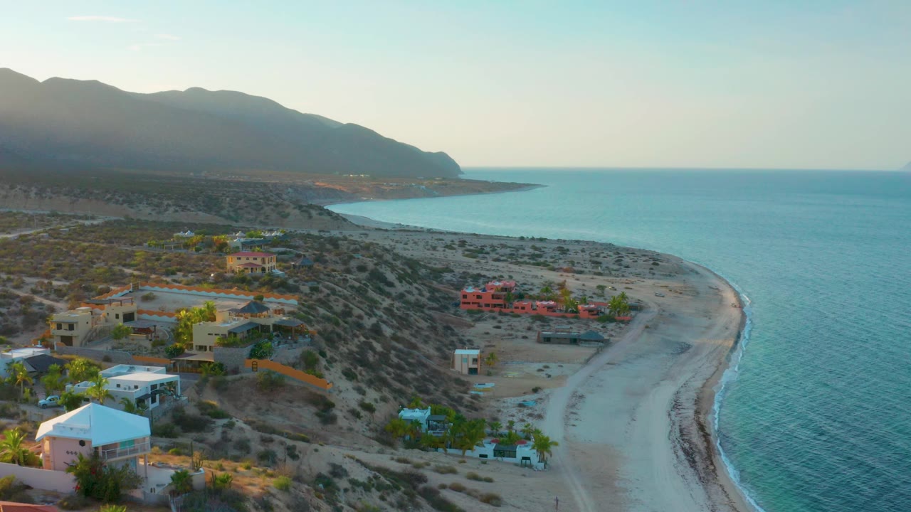 Drone perspective over El Sargento La Ventana, Mexico with turquoise sea on the right, a quiet sandy beach below, small coastal buildings, desert vegetation, mountains, and no visible people.