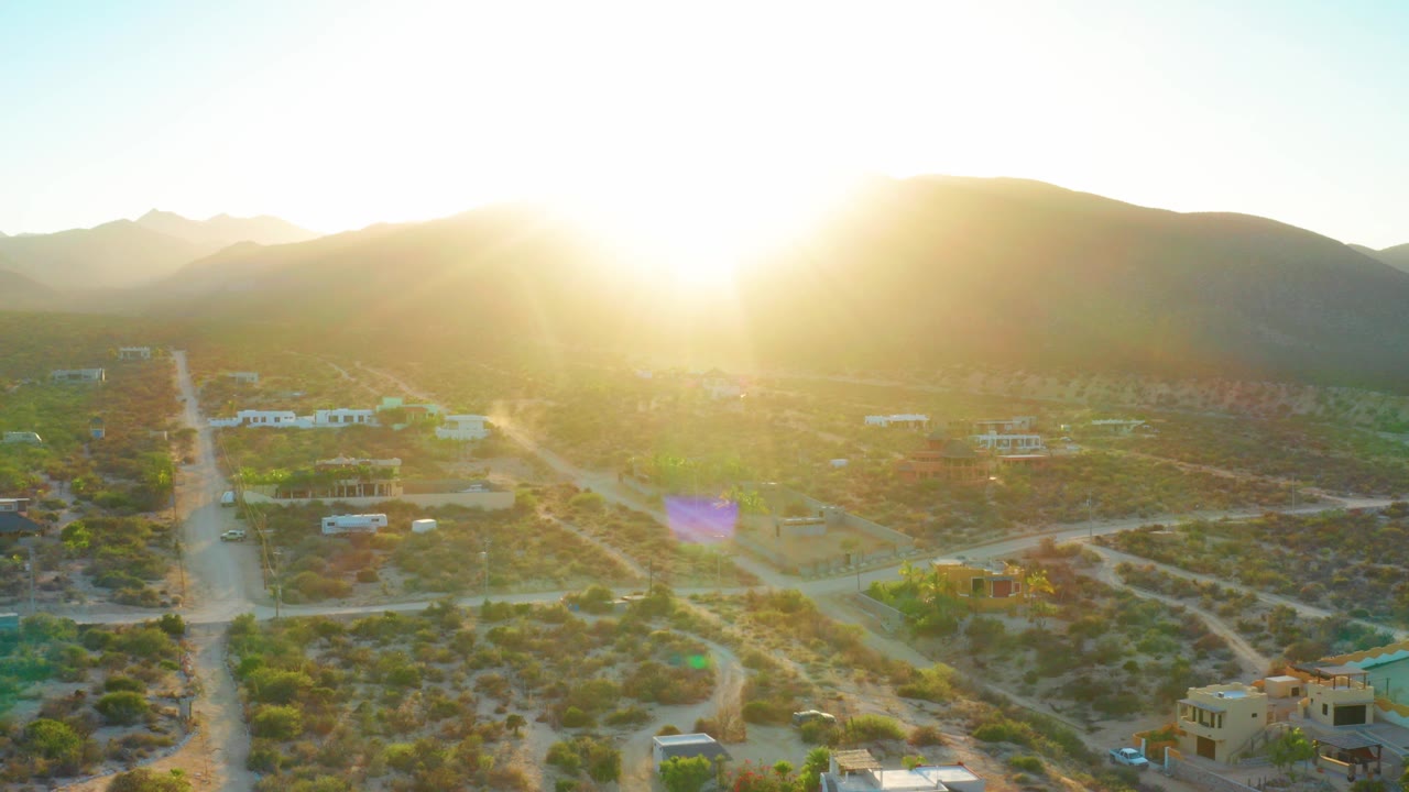 Aerial view of El Sargento La Ventana, Mexico at sunrise, with dirt roads, scattered homes, desert shrubs, hills on the horizon, and no visible people.