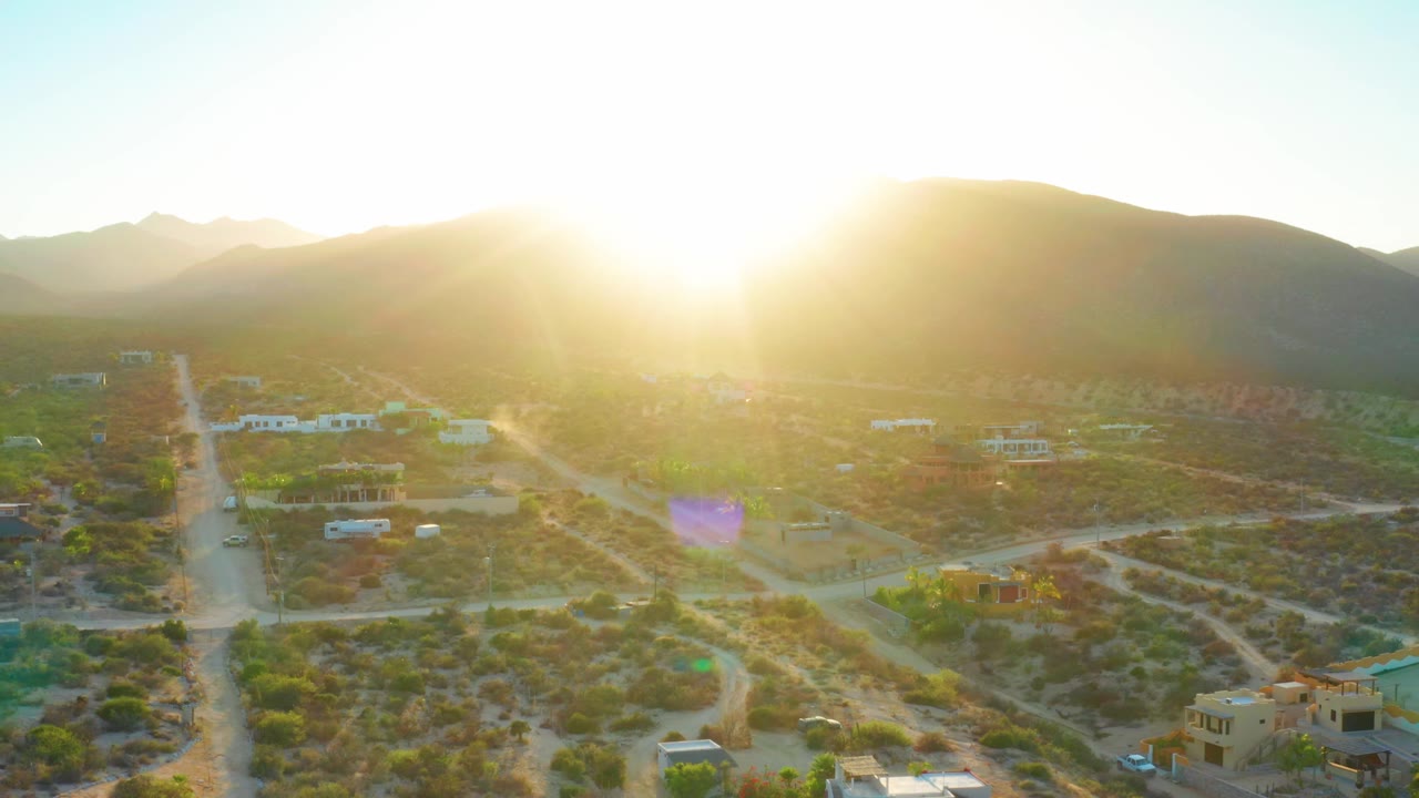 Sun flare hangs above El Sargento La Ventana, Mexico, with a straight dirt road at left, scattered homes below, dry vegetation, hills behind, and no visible people.