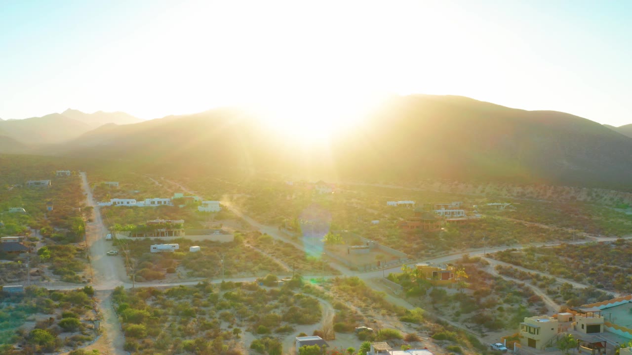 Aerial sunrise frame in El Sargento La Ventana, Mexico, showing dark hills, washed morning light, scrubland lots, several homes, dirt roads, and no visible people.
