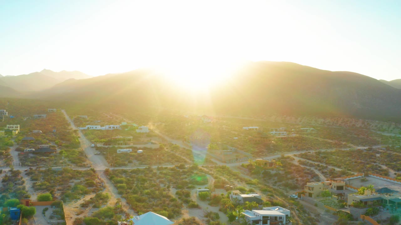 High aerial view over El Sargento La Ventana, Mexico, with low sun above mountain ridges, dirt roads, scattered houses, scrub terrain, and no visible people.