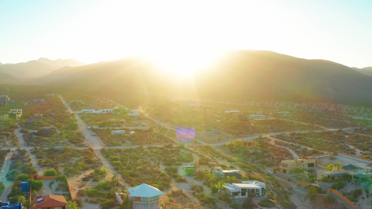 Wide aerial view of El Sargento La Ventana, Mexico, with the sun near the ridge line, a road grid across the valley, scattered buildings, and no visible people.