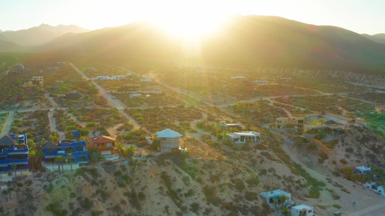 At 0:00 in El Sargento La Ventana, the sun flares over dark mountain ridges above scrub-covered lots, scattered homes, and a strip of beach, with no visible people.