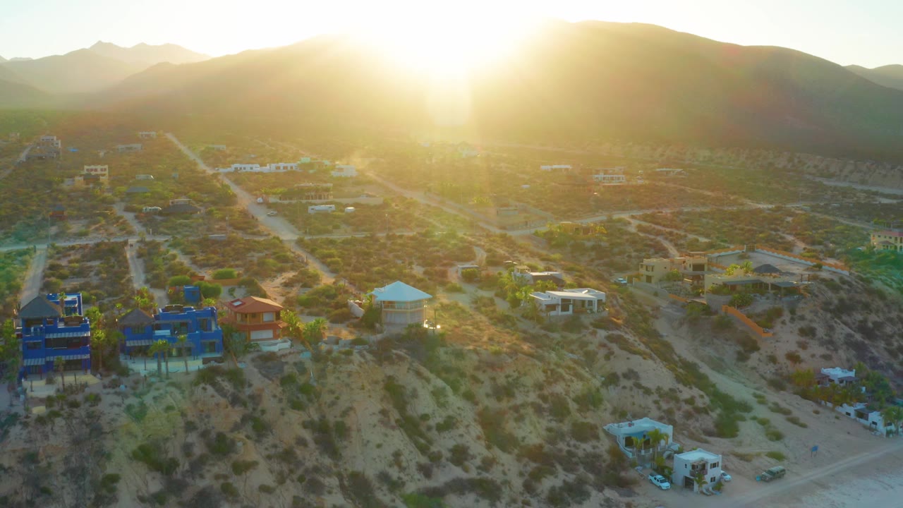 At 0:02, sun glare washes the upper frame while blue and red-roofed homes sit among scrub and roads below the mountains in El Sargento La Ventana, with no visible people.