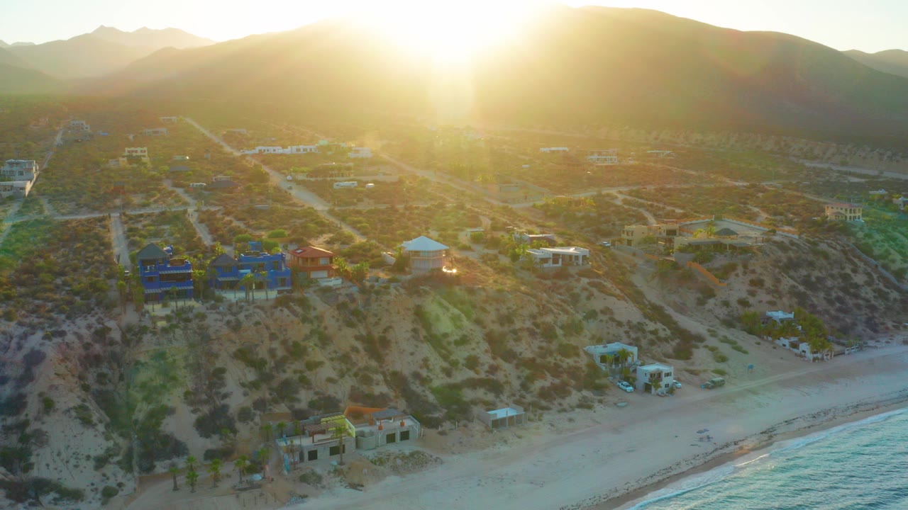 Aerial view in El Sargento La Ventana, Mexico, showing beach at the bottom, hillside homes and dirt roads, mountains behind, intense low sun flare, and no visible people.