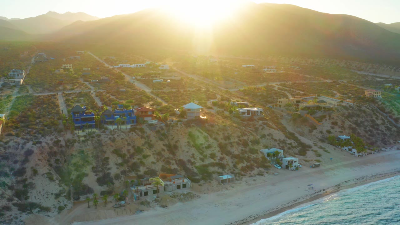 Drone-style aerial view in El Sargento La Ventana, Mexico, showing pale sand, a strip of surf, hillside residences, winding dirt roads, scrub-covered slopes, distant mountains, bright sun, and no visible people.