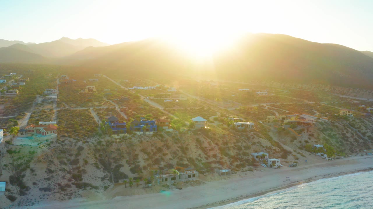 Opening aerial frame over El Sargento La Ventana, Mexico, with the sun flaring above mountains, scattered homes on arid slopes, beach and water below, and no visible people.