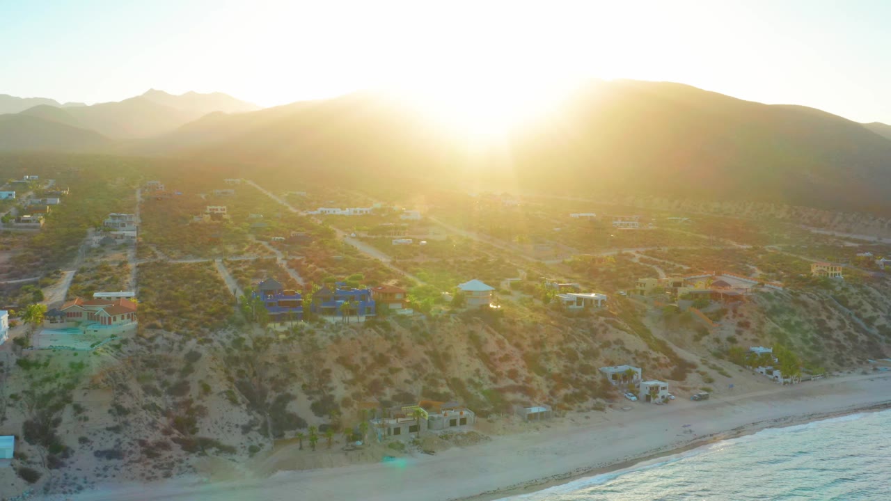 Aerial view in El Sargento La Ventana with bright sunrise glare near the ridge, dirt roads crossing the hillside, small houses above the beach, and no visible people.