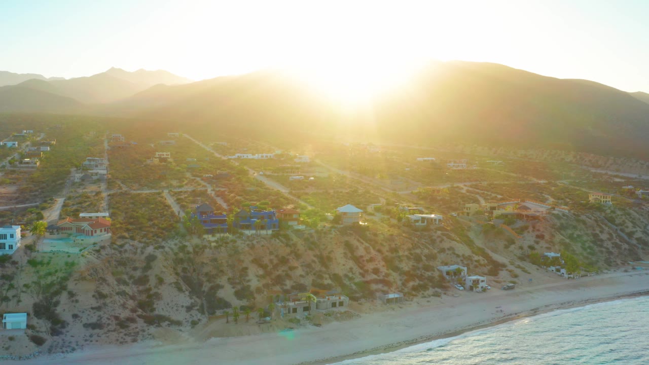 The sun blooms over the mountain horizon in El Sargento La Ventana while houses, graded roads, sandy beach, and a strip of ocean remain visible with no visible people.