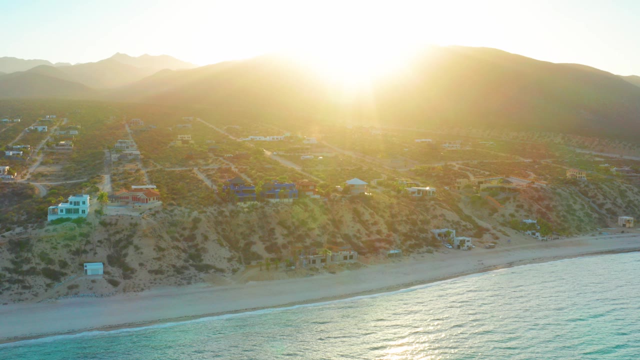 At 0:00, the sun sits just above the hills in El Sargento La Ventana, Mexico, with turquoise water in front, beach and small coastal buildings behind it, and no visible people.