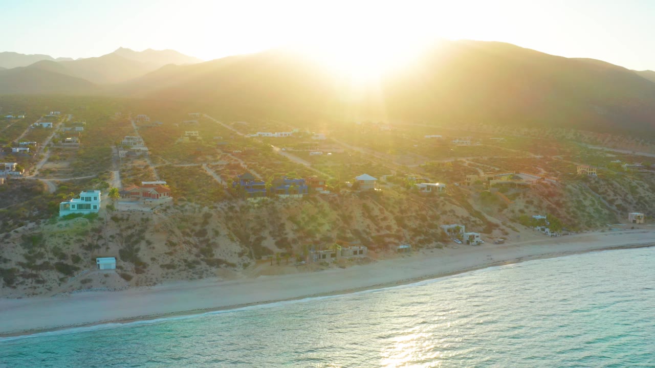 At 0:02, the aerial view holds turquoise water and a thin beach below hillside roads and scattered buildings in El Sargento La Ventana, Mexico, with the sun glaring near the top edge and no visible people.