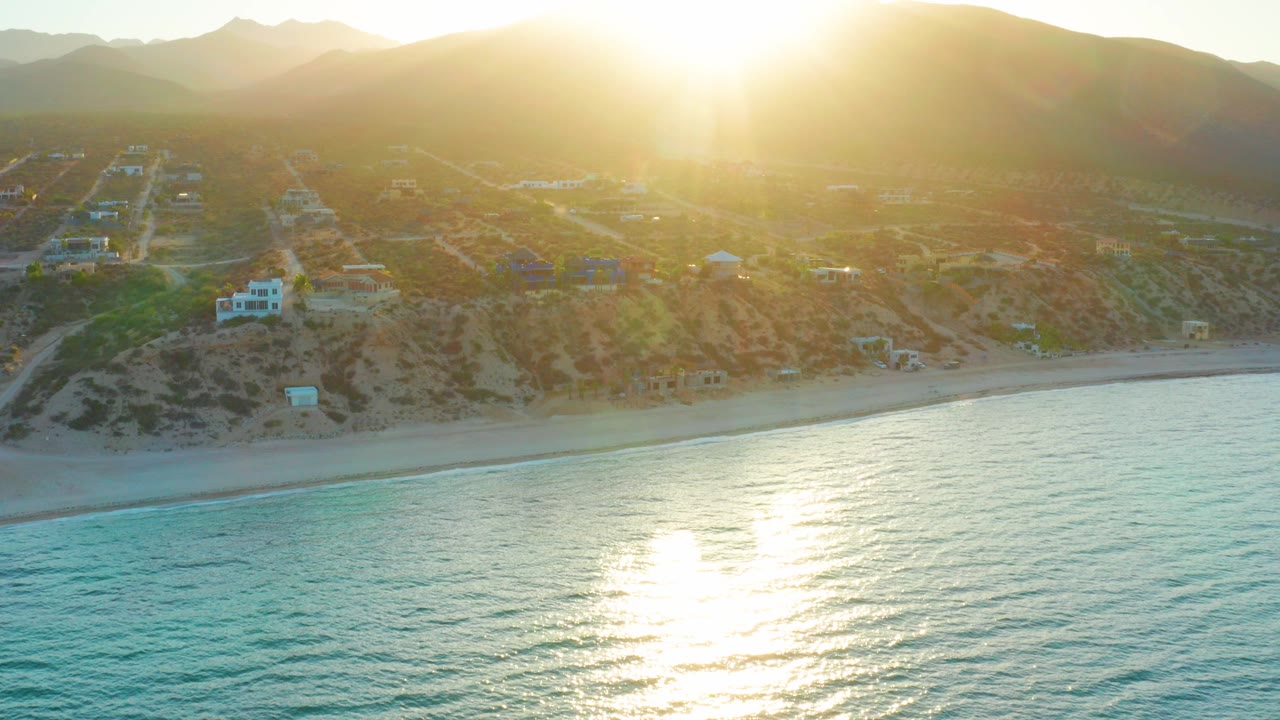 Aerial view of El Sargento La Ventana, Mexico, with bright sun glare on turquoise water, a sandy beach, hillside homes, and no visible people.