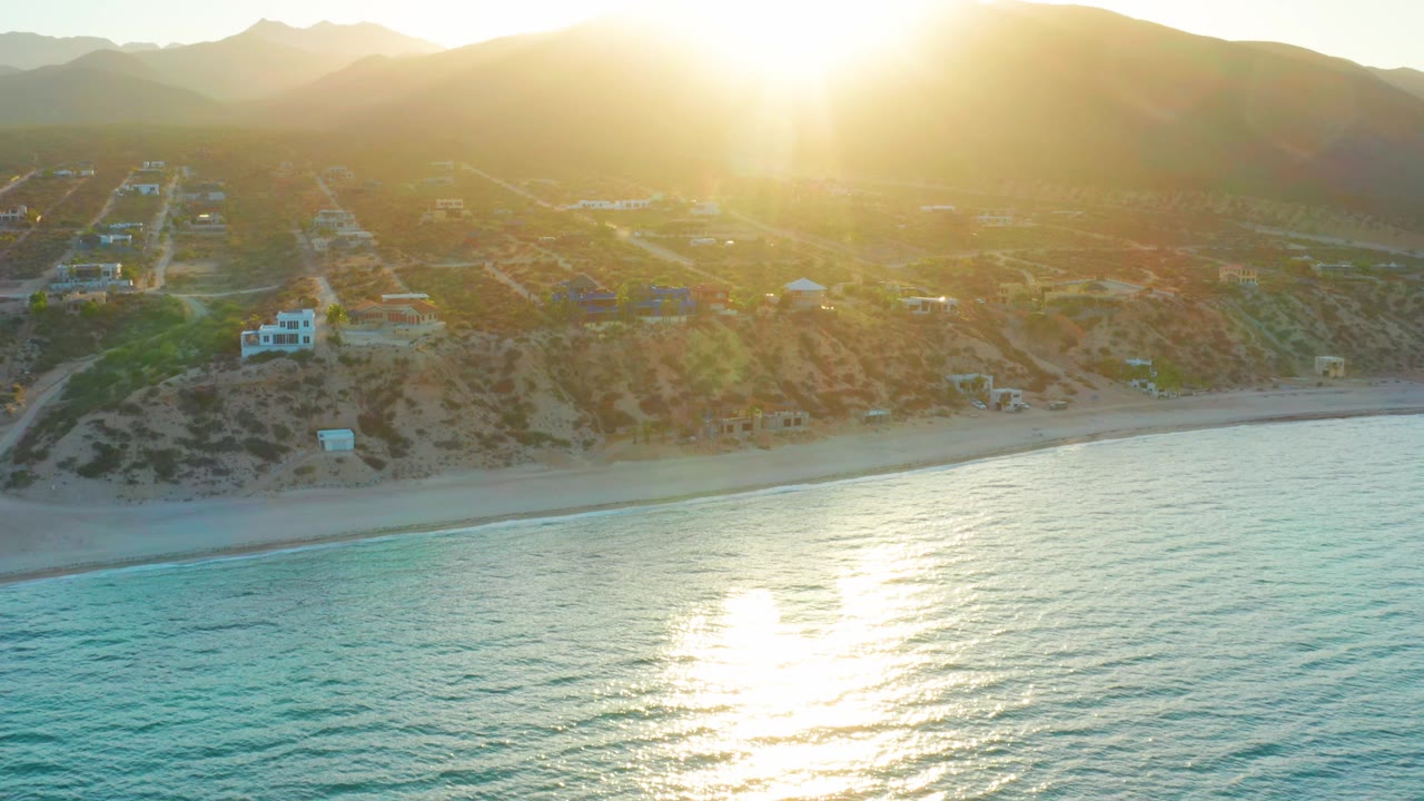 Aerial coastal frame in El Sargento La Ventana showing intense backlight, rippled water, a narrow beach, hillside roads, homes, and no visible people.
