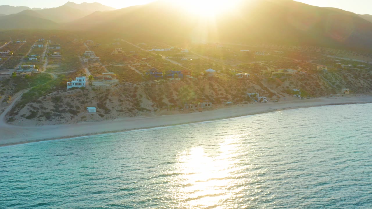 Wide aerial view of El Sargento La Ventana with the ocean filling the foreground, sun flare on the surface, beach and hillside homes in back, and no visible people.
