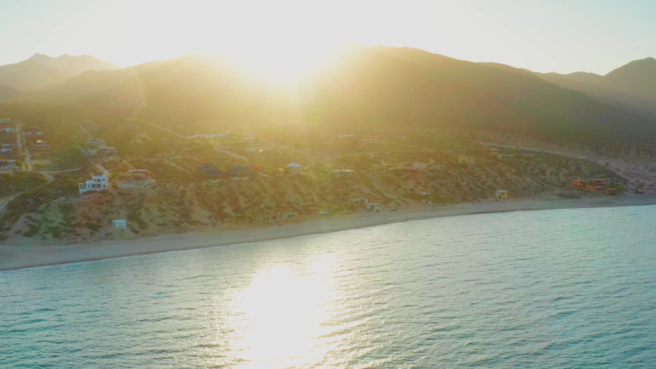 Opening frame of El Sargento La Ventana, Mexico, with the sun breaking over the mountain horizon, ocean in the foreground, hillside homes near the beach, and no visible people.