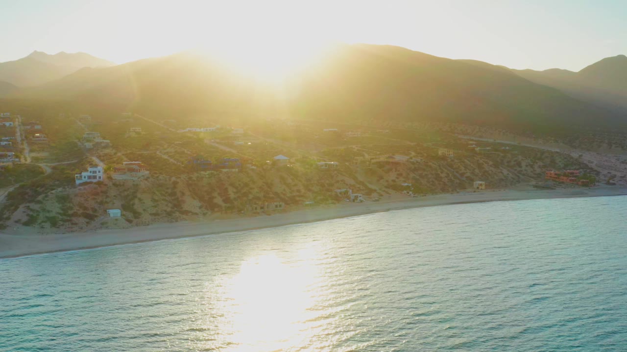 Early frame of El Sargento La Ventana, Mexico, showing intense sun flare over the ridgeline, blue-green water below, a thin beach strip, scattered homes, and no visible people.