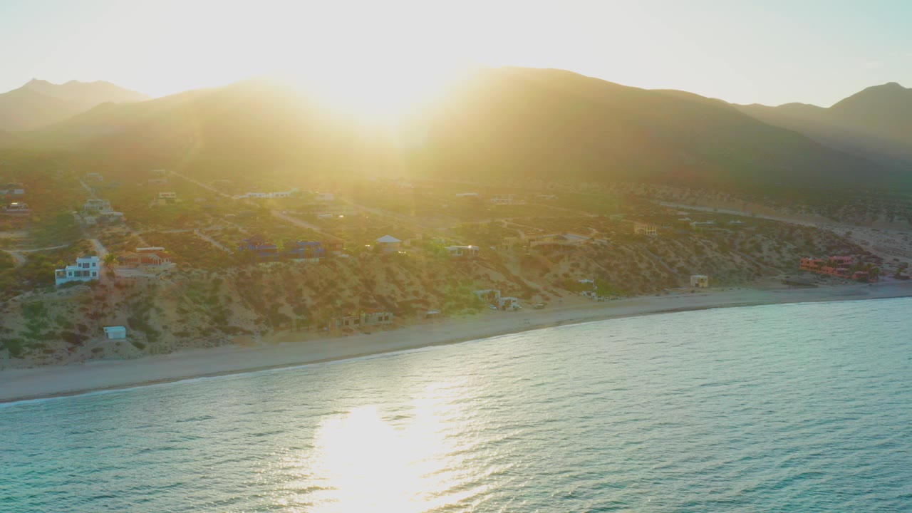 Opening aerial frame over El Sargento La Ventana, Mexico, with turquoise water below, a narrow beach and hillside settlement ahead, bright low sun over the mountains, and no visible people.