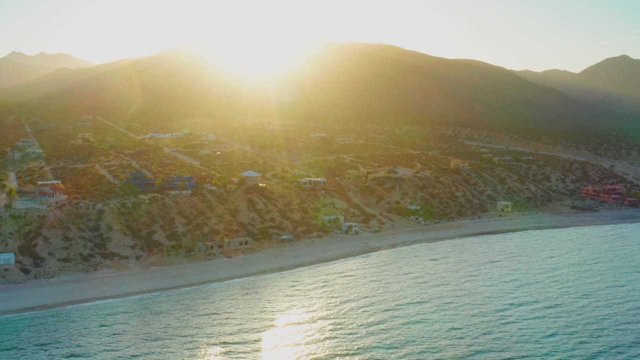 Opening aerial view of El Sargento La Ventana, Mexico with blue-green water, sandy beach, hillside homes, mountains, strong sun flare, and no visible people.