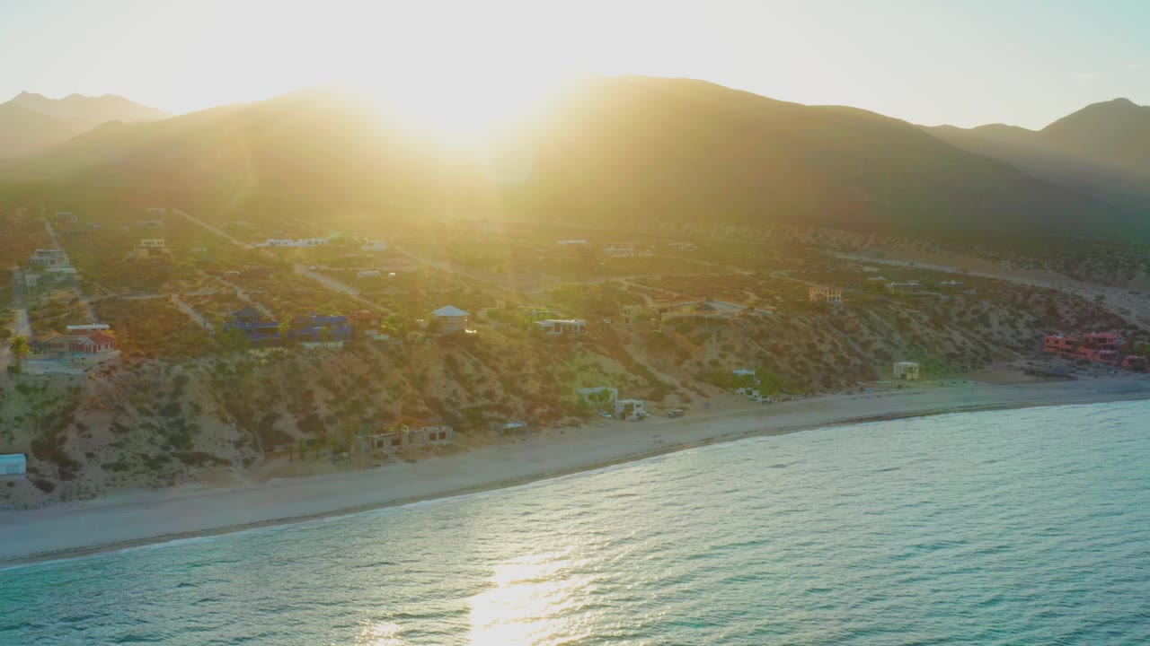 Aerial frame of El Sargento La Ventana shoreline where the sun reflection crosses the water below beach houses and a dark mountain ridge, with no visible people.