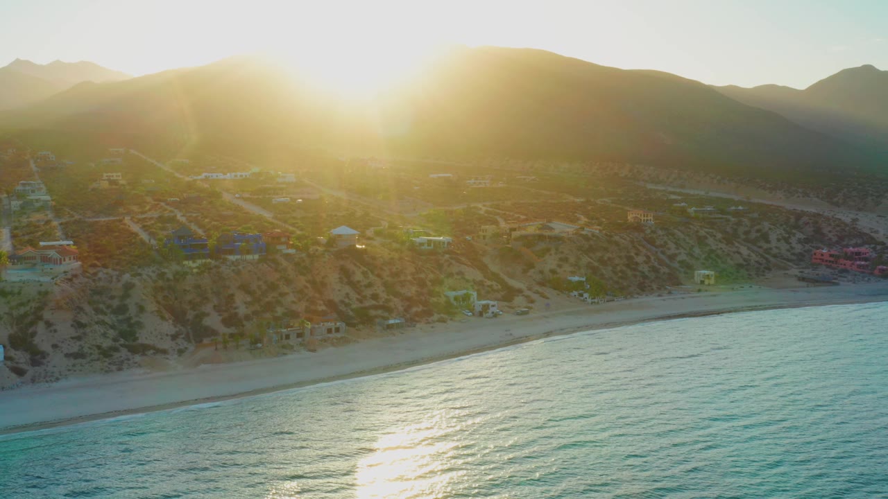 Aerial view in El Sargento La Ventana, Mexico with the sun centered over the hills, beach houses along the sand, textured blue-green water below, and no visible people.
