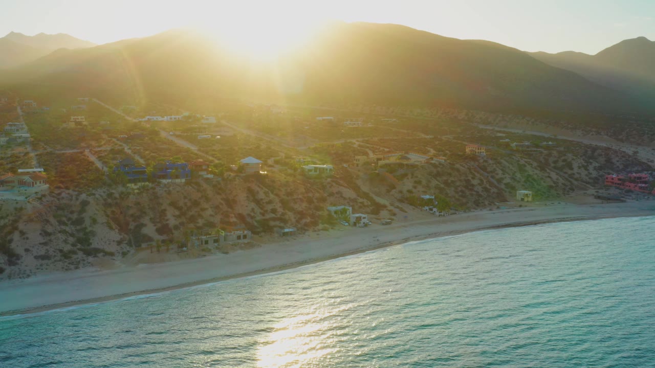 Aerial coastal hillside with strong sun flare and waves along a sandy beach.