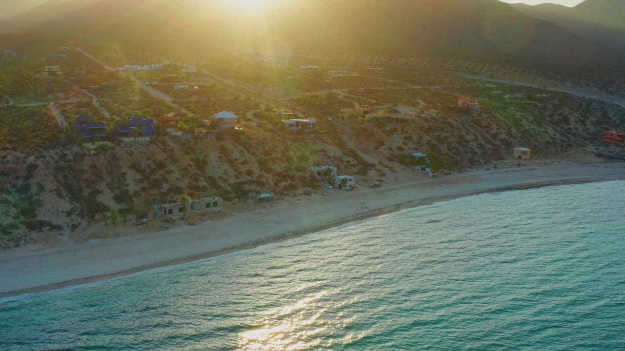 Aerial view in El Sargento La Ventana, Mexico with the low sun flaring over hills, turquoise water in the foreground, a sandy beach, scattered coastal homes, and no visible people.