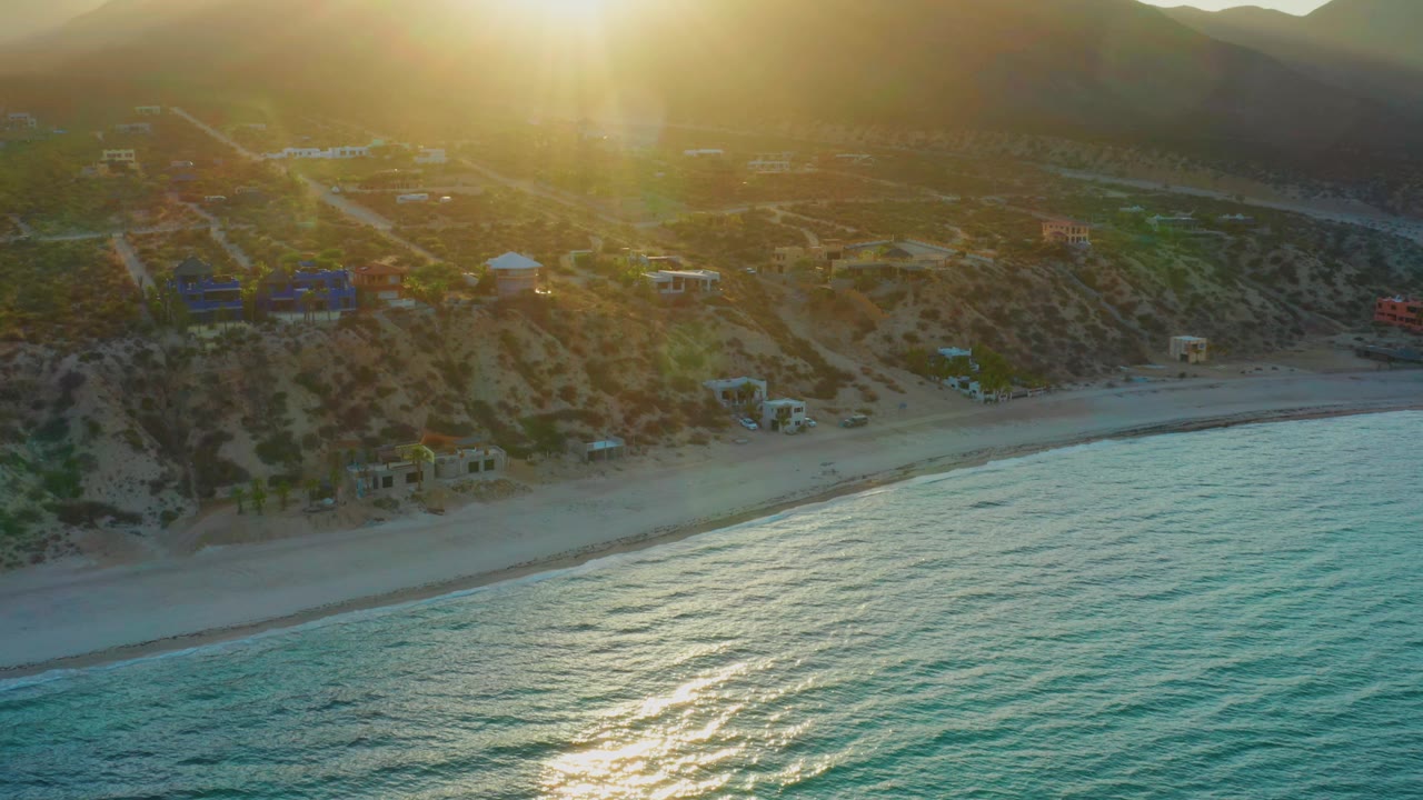 Wide aerial frame of El Sargento La Ventana with bright evening glare near the top, rippled blue-green water below, a narrow beach, hillside roads, buildings, and no visible people.
