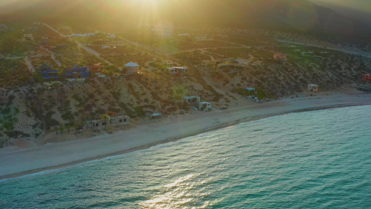 Aerial sunset view in El Sargento La Ventana showing the bright sun above hazy hills, open ocean across the lower frame, a pale beach line, small buildings, and no visible people.