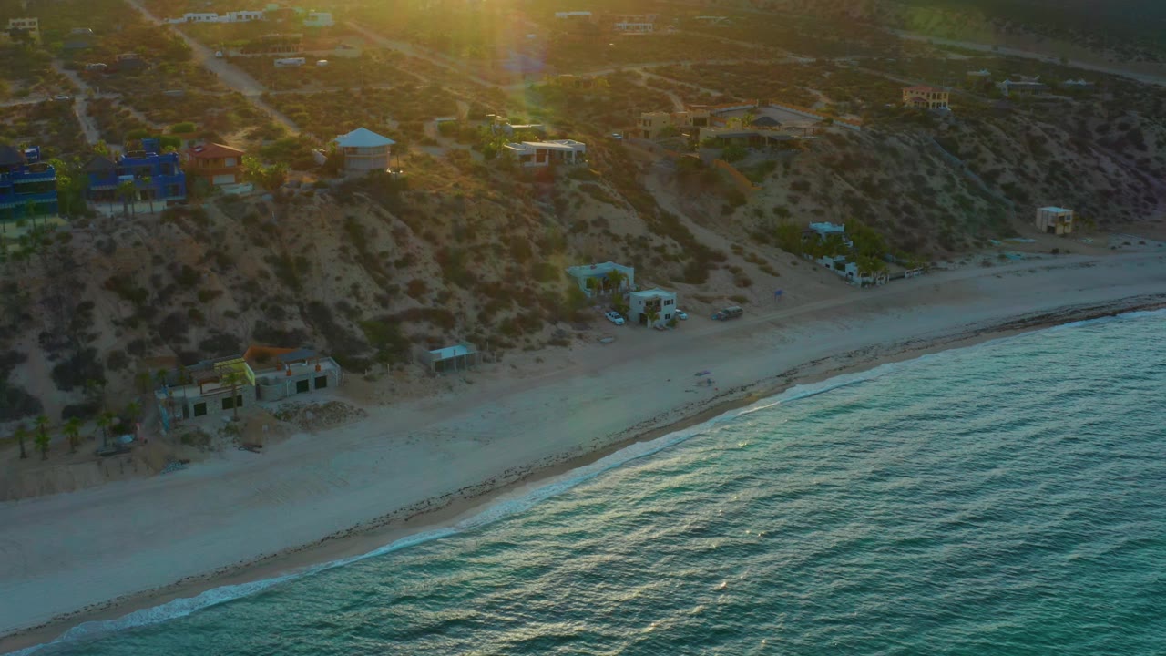 Aerial view in El Sargento La Ventana, Mexico, with the surf line cutting across the lower frame, sandy beach below hillside roads and scattered structures, and no visible people.